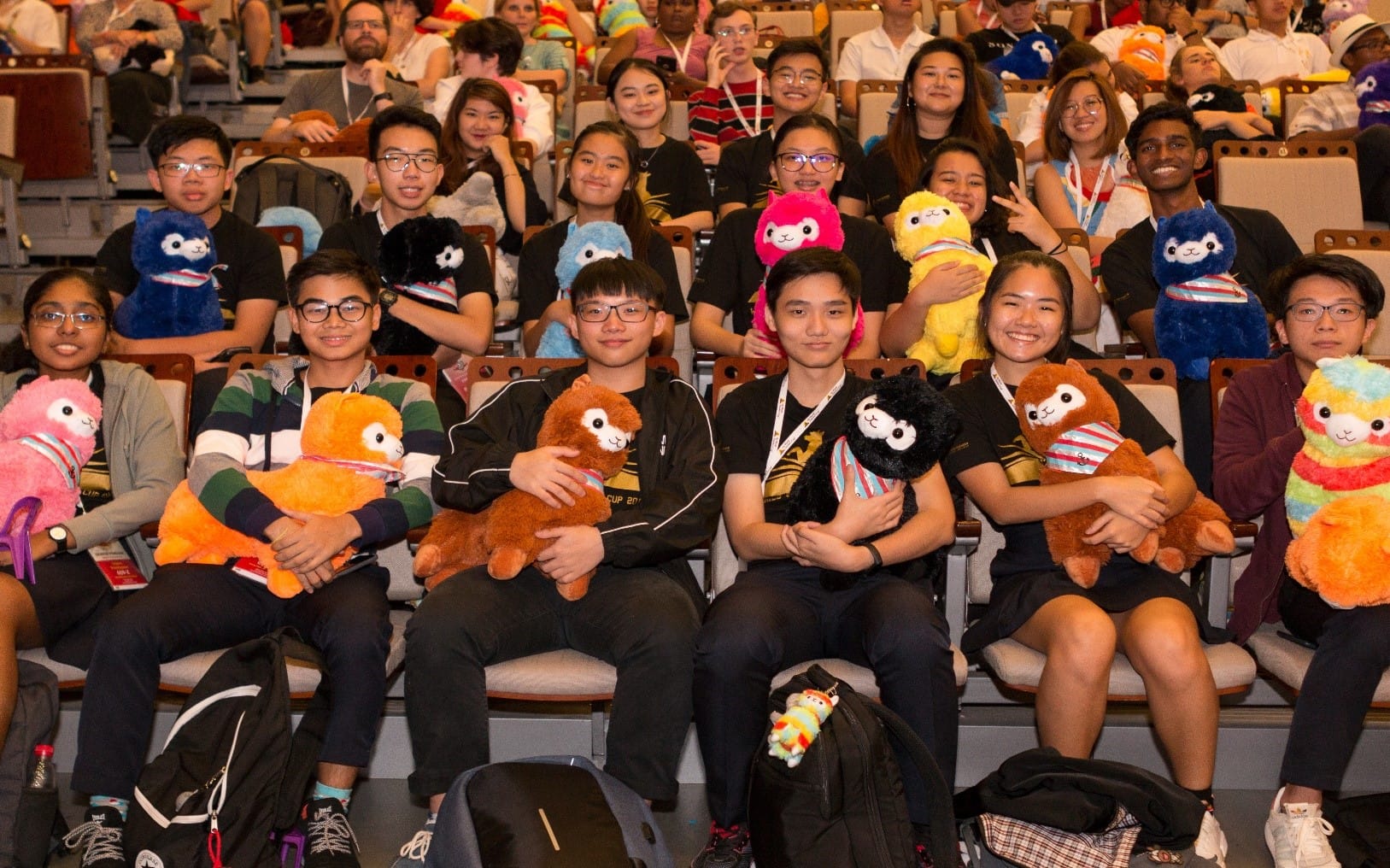 Young people seated in an auditorium hold colorful stuffed alpaca plush toys and smile at the camera.